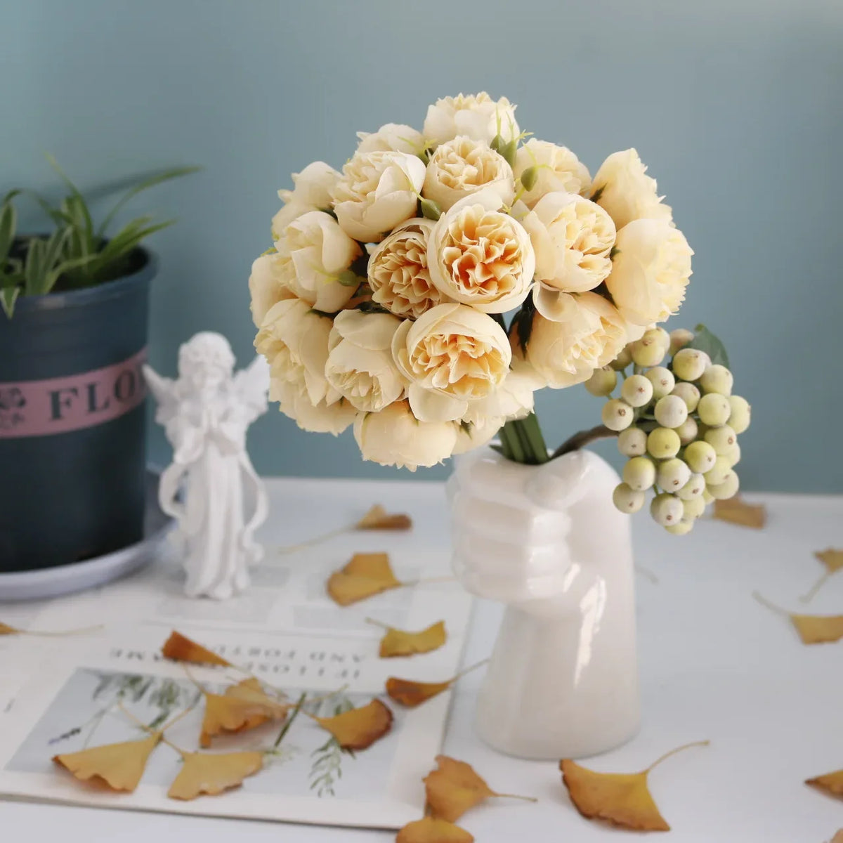 A bouquet of cream-colored artificial peony silk flowers in a white vase on a white surface.