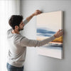 A man is hanging a large framed canvas wall art featuring a sunset and blue sky over the ocean.