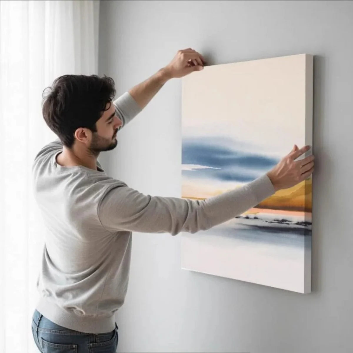 A man is hanging a large framed canvas wall art featuring a sunset and blue sky over the ocean.