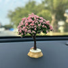 A small tree ornament with pink flowers and a trunk sits on a dark dashboard surface.
