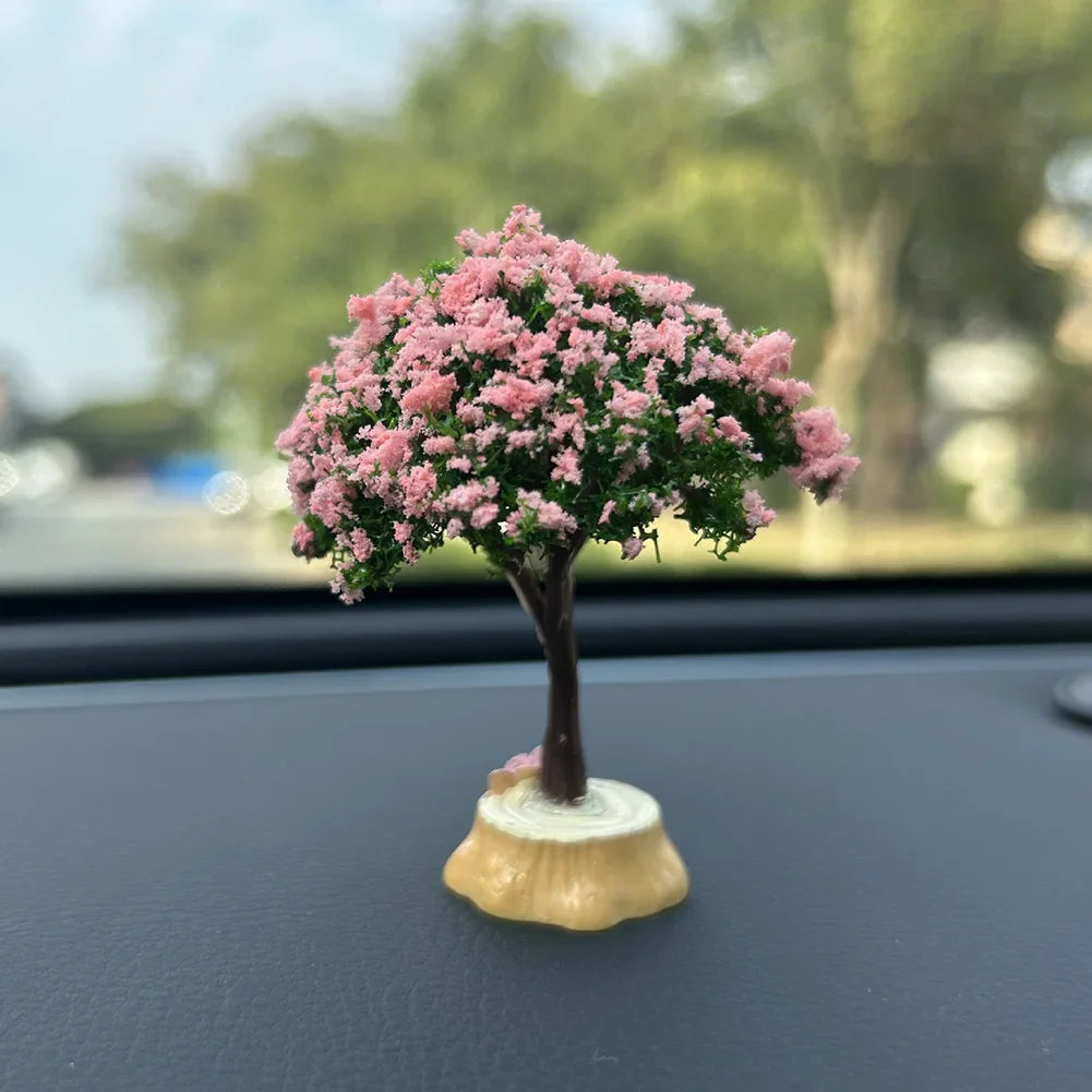 A small tree ornament with pink flowers and a trunk sits on a dark dashboard surface.