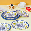 A stack of blue floral paper plates and a plate with a red cake are displayed on a light yellow surface.