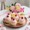 A display of pink and gold elegant crown and bow cupcake toppers on a wooden platter.