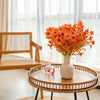 An artificial autumn bouquet of vibrant orange and yellow silk flowers displayed in a white vase on a wooden table.