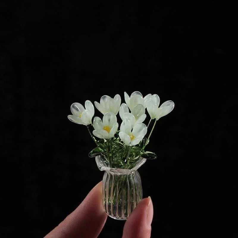 A hand holds a small glass vase filled with white handcrafted mini flower sculptures against a black background.