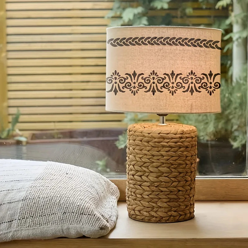 A woven cylindrical lamp shade with a decorative braided rim sits on a wooden surface next to a light-colored pillow.