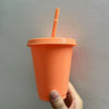 An orange plastic cold cup with a lid and straw is being held against a light gray wall.