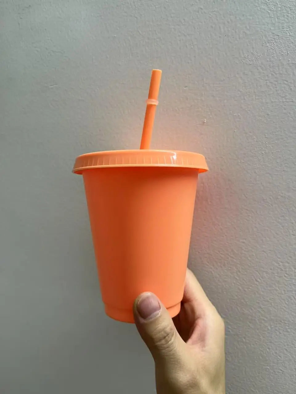 An orange plastic cold cup with a lid and straw is being held against a light gray wall.