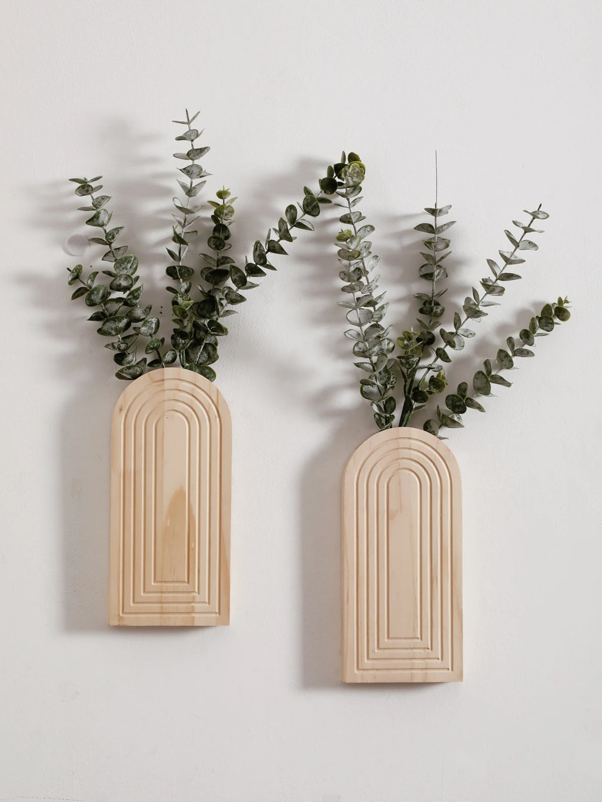 Dos jarrones de pared arqueados de madera contienen follaje verde seco contra una pared blanca.
