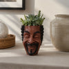 A terracotta planter shaped like a smiling face holds a succulent plant on a light-colored table.