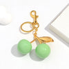 A gold-colored resin pendant keychain featuring a pink cherry charm and two light green spherical beads.