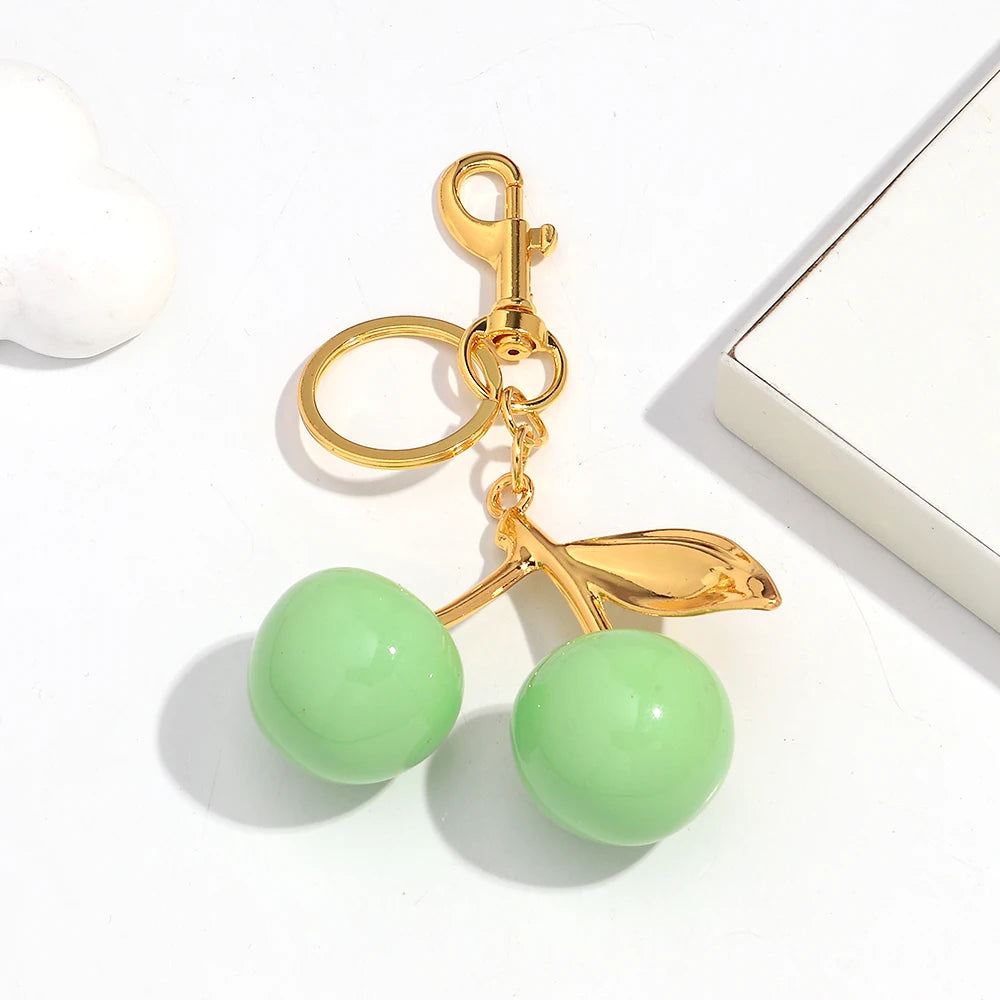 A gold-colored resin pendant keychain featuring a pink cherry charm and two light green spherical beads.