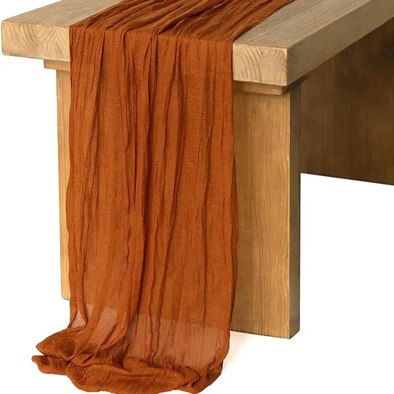 A rust orange gauze table runner draped over a light wood table.