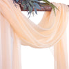 Light peach chiffon fabric panels draped over a wooden arch with green foliage.