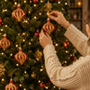 A person is hanging a red and gold Christmas ornament on a decorated green Christmas tree.
