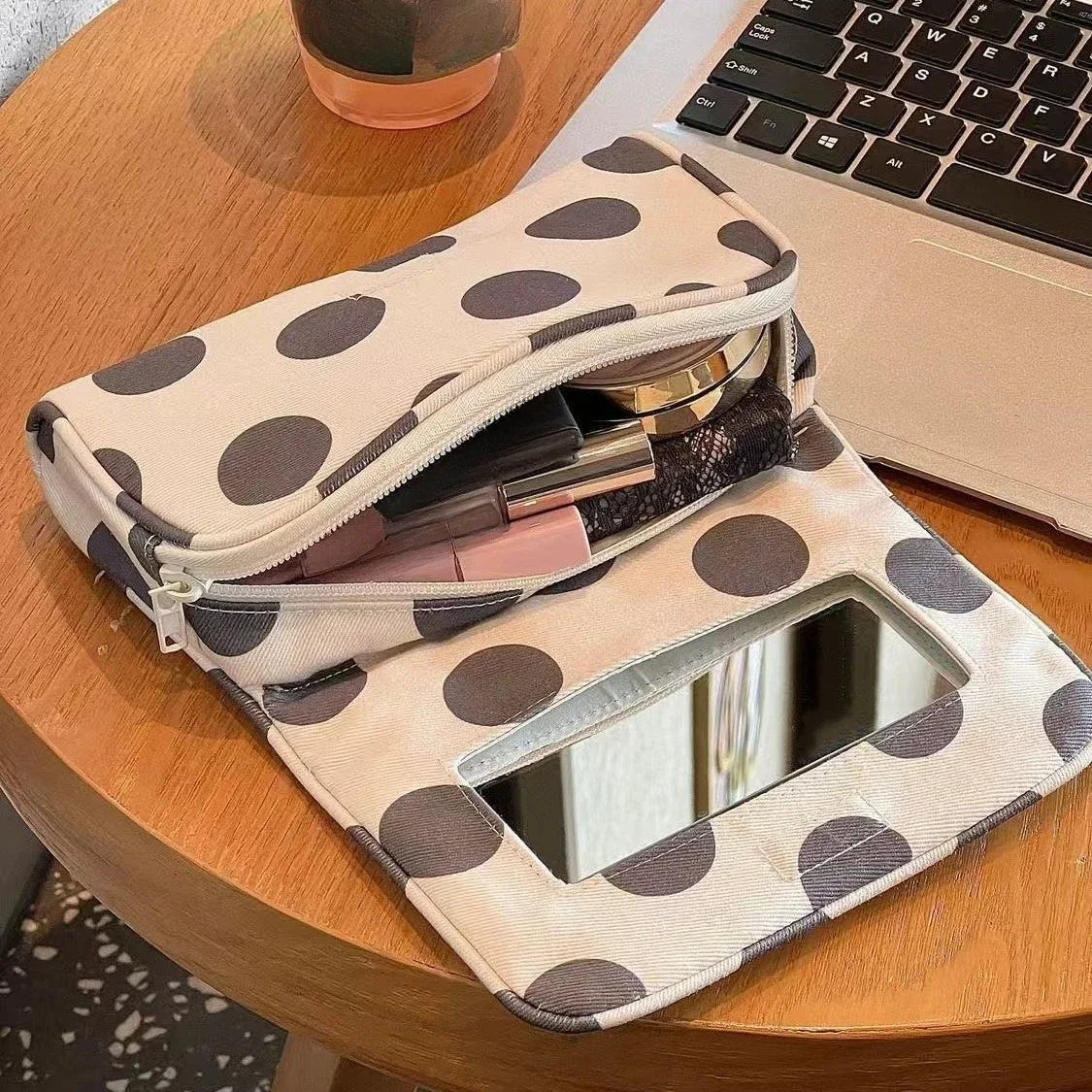A beige canvas travel makeup bag with polka dots and a mirror is resting on a wooden table next to a laptop.