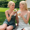 Two blonde women smiling while holding disposable plastic cups with lids and straws outdoors