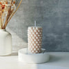 A white candle with a textured surface of small bubbles sits on a white dish next to dried floral stems.