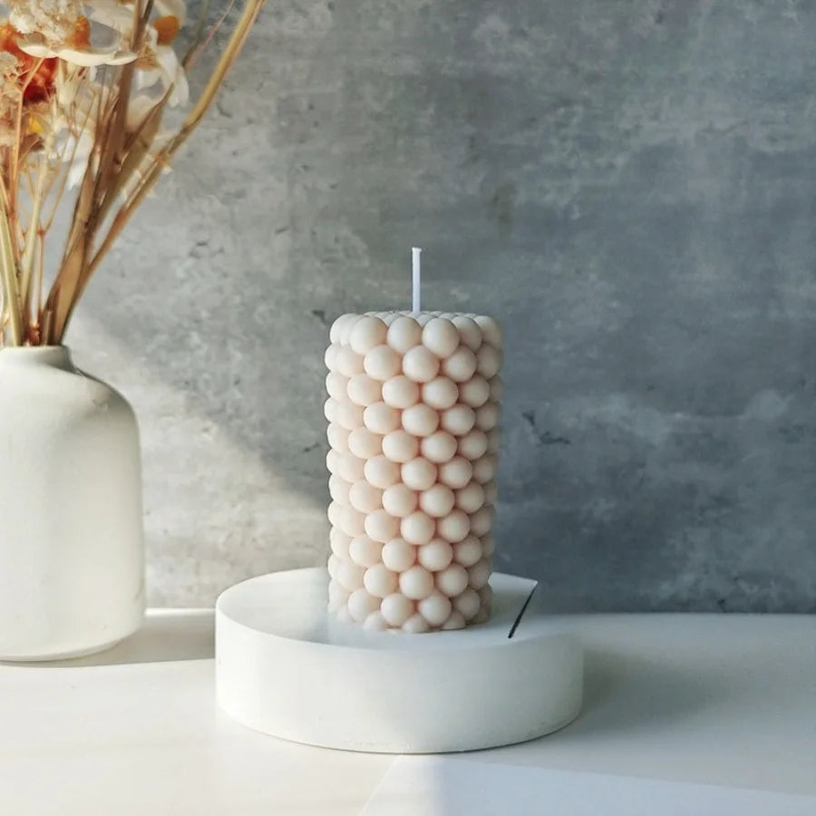 A white candle with a textured surface of small bubbles sits on a white dish next to dried floral stems.