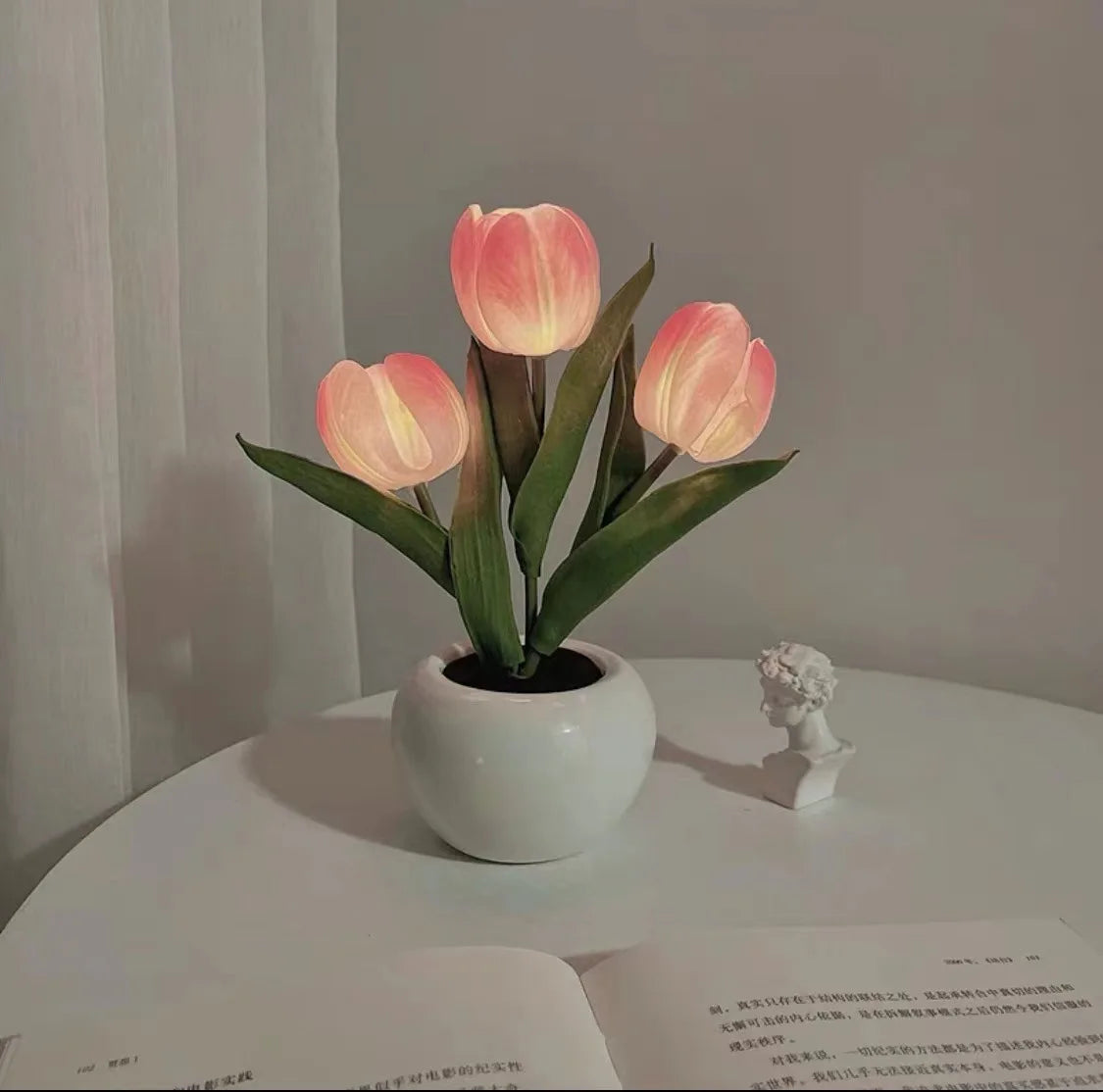 A pink LED tulip night light with petals placed in a white ceramic pot on a table.