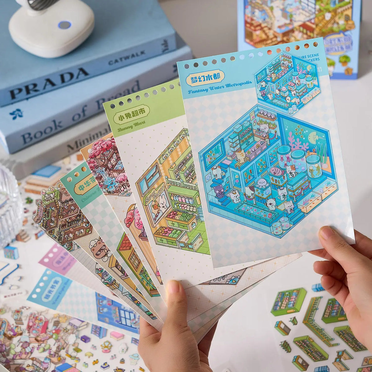 A hand holds a colorful sticker book featuring kawaii miniature 3D scenes and buildings for kids.