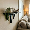 A black drip floating shelf displaying potted greenery and a small brown bird figurine on a light-colored wall.