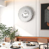 A modern silver wall clock with a white face and black numbers is mounted on a light gray wall in a contemporary interior.