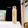 A clear glass bottle filled with yellow Citrine crystals sits on a white surface.