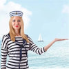 A blonde woman wearing a striped shirt and a captain's hat stands by the sea.