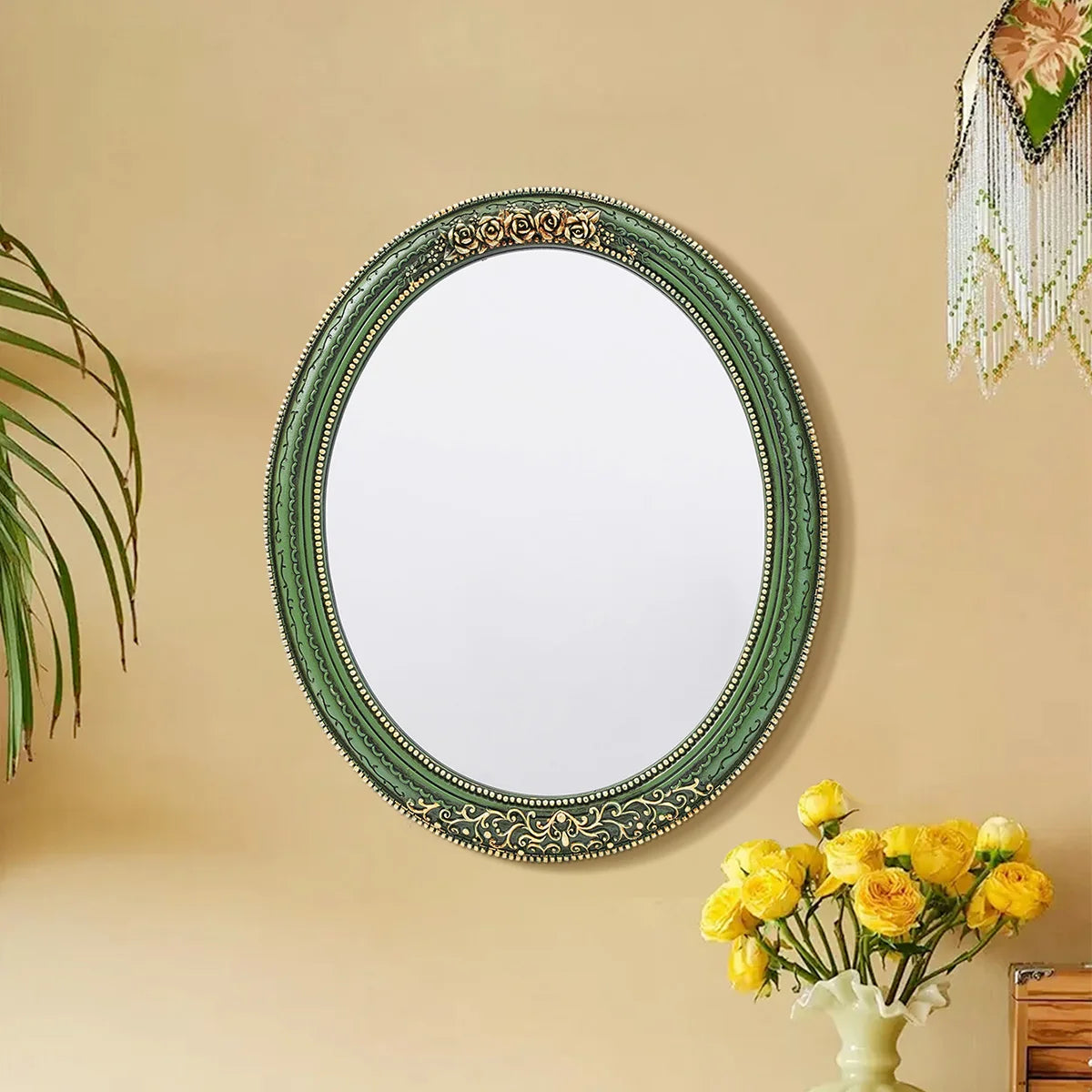 An oval wall mirror with a green frame and intricate lace design hanging on a beige wall.