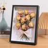A black shadow box frame holds a dried floral arrangement with pink and white accents.