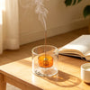 A gourd-shaped glass incense burner with an orange-colored stick emitting smoke on a wooden table.