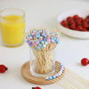 A glass jar of yellow juice next to a glass container filled with colorful beaded bamboo fruit skewers on a wooden coaster.