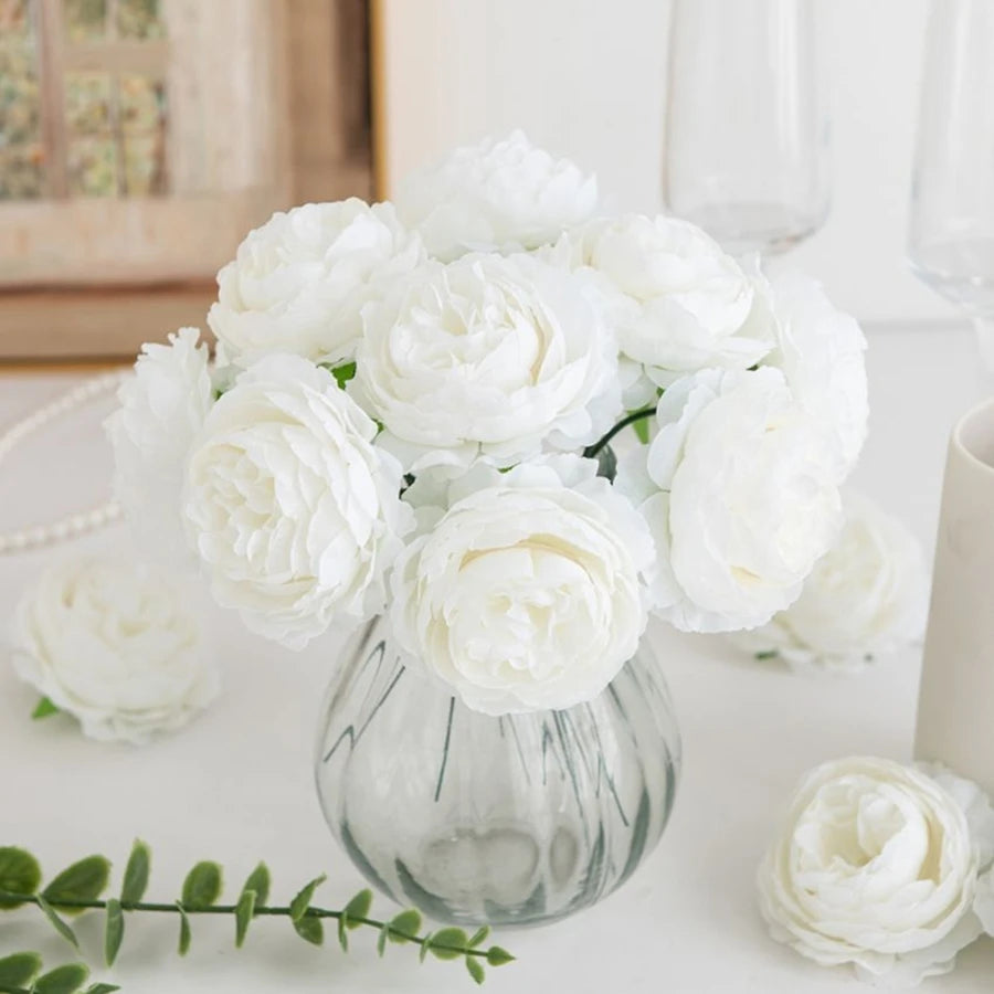 A bouquet of white silk peony artificial flowers in a clear glass vase on a white surface.