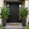A realistic faux areca palm tree in a black planter stands on a stone step in front of a dark door.