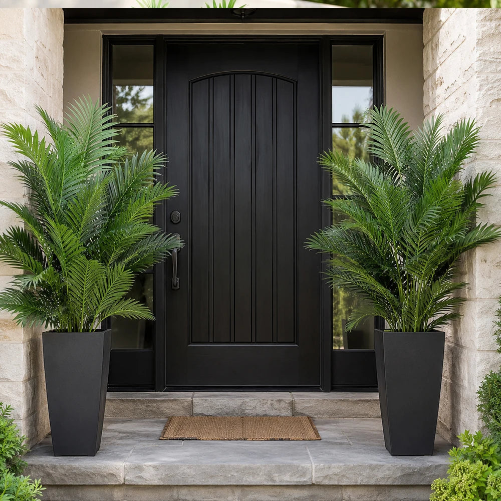 A realistic faux areca palm tree in a black planter stands on a stone step in front of a dark door.