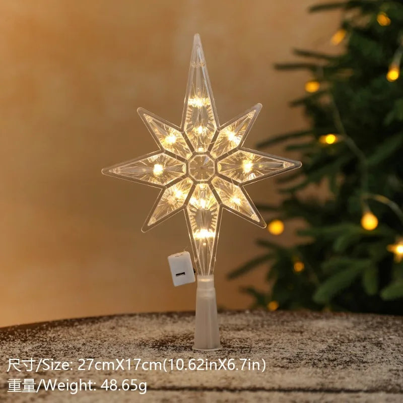 A clear LED Christmas tree star topper with warm white lights displayed on a wooden surface next to a green decorated tree.