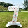 A white standing foil balloon shaped like the number one with a crown and birthday text, placed on green grass.