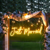 A glowing neon sign spelling Just Married is displayed on a wooden arch decorated with flowers and draped fabric.