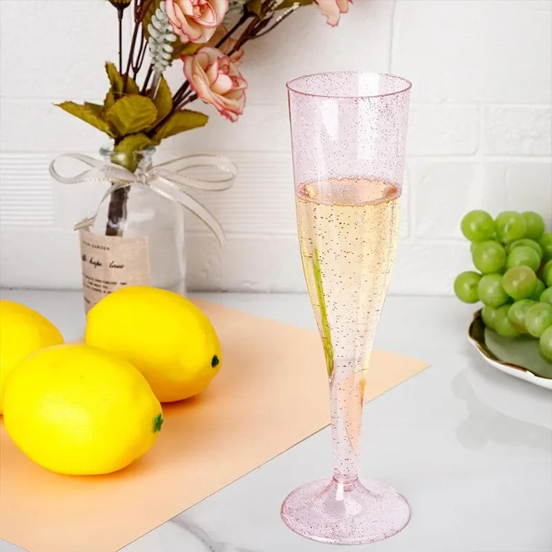 A tall pink glitter disposable champagne flute stands next to a yellow lemon and a bunch of green grapes.