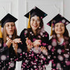 Three young women in graduation gowns smiling and throwing pink confetti dots into the air