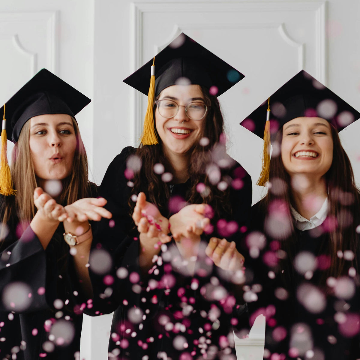 Three young women in graduation gowns smiling and throwing pink confetti dots into the air