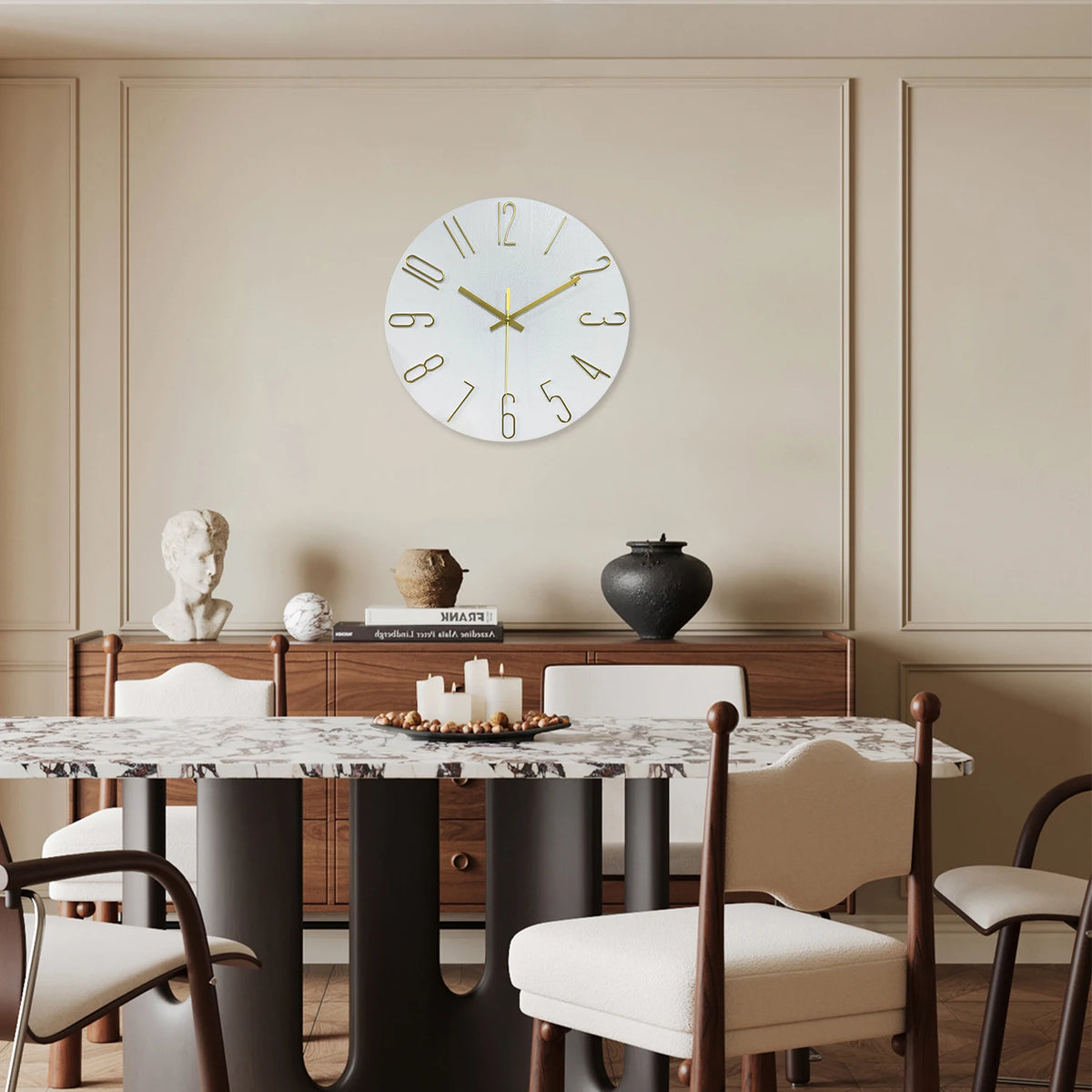 A round modern wall clock with gold 3D numerals hangs on a light beige wall above a wooden cabinet.