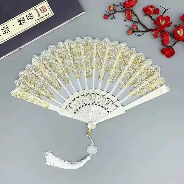 A white and gold folding fan with delicate lacework and red floral accents displayed on a gray surface.