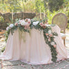A floral centerpiece featuring pink and white roses and eucalyptus garlands on a draped table for an event.