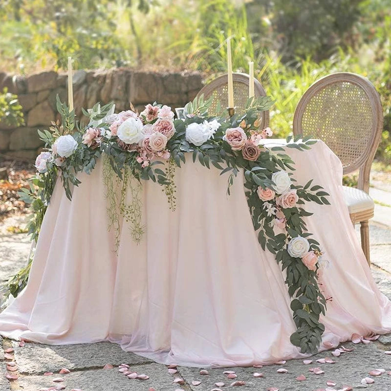 A floral centerpiece featuring pink and white roses and eucalyptus garlands on a draped table for an event.