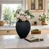 A black ceramic vase holds a bouquet of dusty pink and green flowers on a white marble countertop.