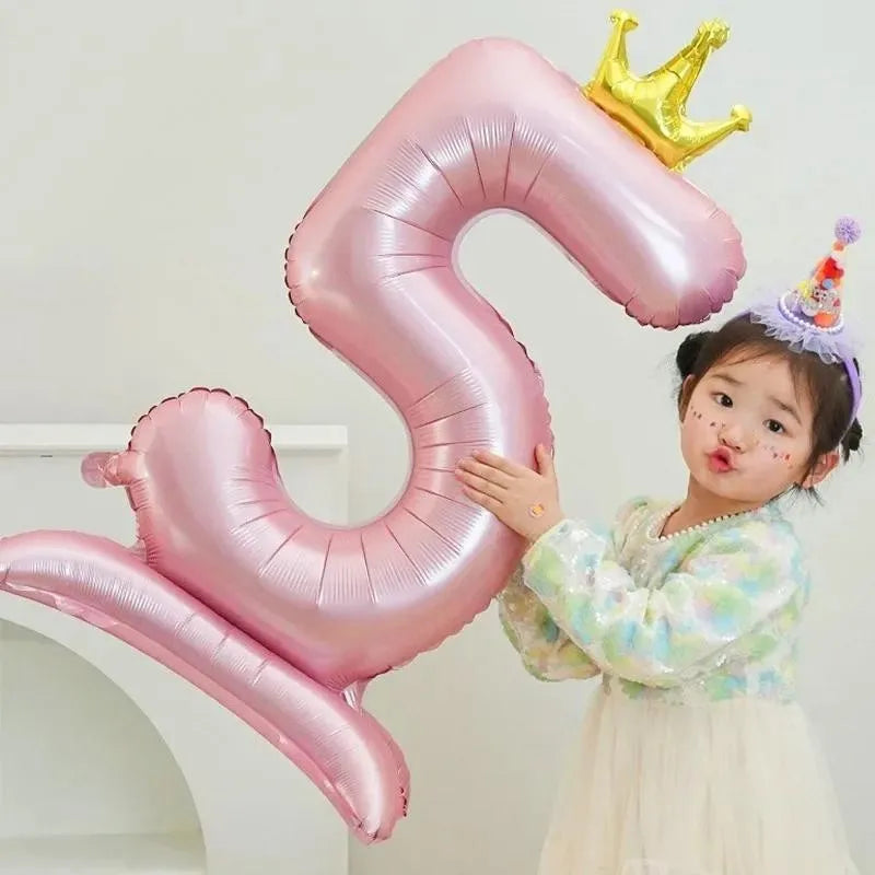 A young girl is posing with a large pink foil balloon shaped like the number 9 and a gold crown.