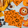 Orange paper napkins featuring black Halloween designs like skulls, spiders, and jack-o'-lanterns on a light wooden surface.