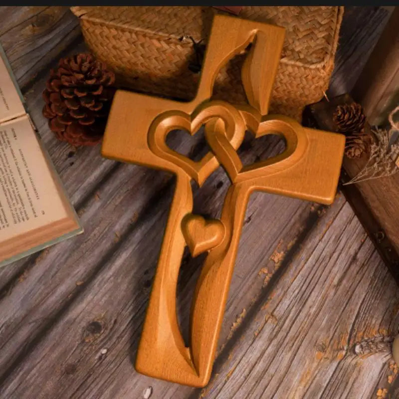 A wooden wall cross with intertwined hearts rests on a rustic wooden surface next to a pinecone and a woven basket.